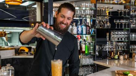 Barman préparant un cocktail en versant une boisson depuis un shaker, devant un bar rempli de bouteilles et de verrerie.