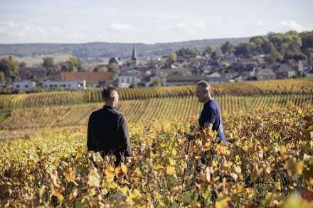 Deux personnes dans un vignoble aux couleurs automnales avec vue sur collines et village en arrière-plan – Fayet Hospitality