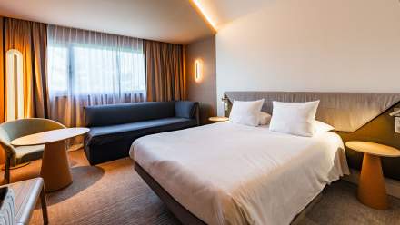 Chambre moderne du Novotel Reims Tinqueux avec grand lit double, canapé gris près de la fenêtre, fauteuil et table ronde, éclairages design et ambiance chaleureuse.