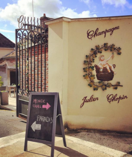 Portail en fer forgé ouvert donnant sur une cour avec mur décoré du logo Champagne Julien Chopin et panneau directionnel indiquant Monde cruel et Paradis - Fayet Hospitality