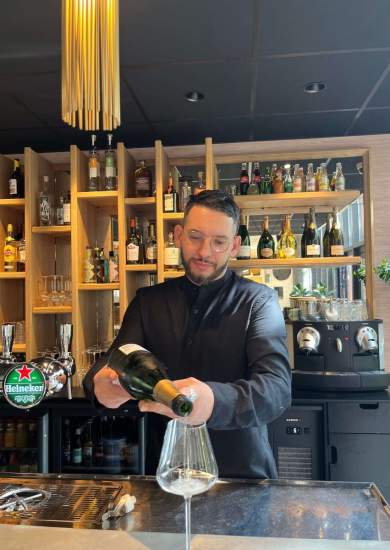 Serveur derrière un comptoir de bar en train de verser du vin dans un verre, étagères remplies de bouteilles en arrière-plan, Fayet Hospitality.