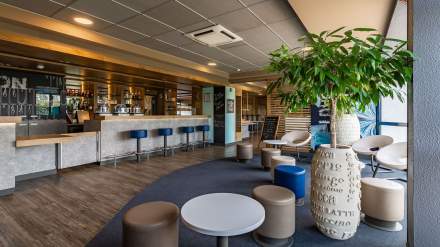 Espace lounge et bar de l’Hôtel Ibis Budget Pontault-Combault, avec comptoir moderne, sièges colorés, tables basses et grande plante décorative.