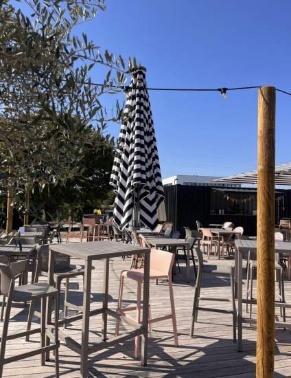 Terrasse en bois avec tables hautes, chaises modernes et parasol rayé noir et blanc sous ciel bleu, Fayet Hospitality.