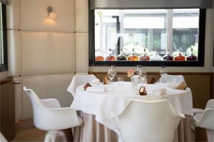 Table élégante dressée avec nappes blanches, verres à pied et assiettes, dans un restaurant lumineux avec vue sur bouteilles en arrière-plan – Fayet Hospitality