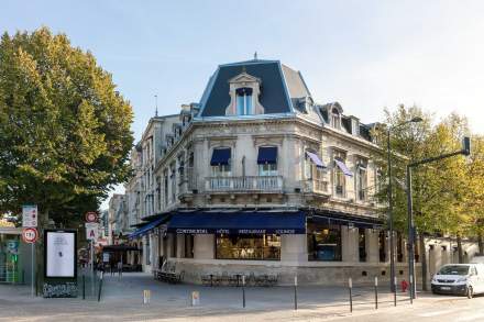 Bâtiment haussmannien élégant avec façade en pierre, fenêtres ornées de balcons et stores bleus, situé à un angle de rue dans un centre-ville animé – Fayet Hospitality