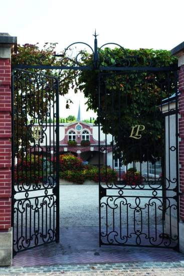 Portail en fer forgé ouvert donnant sur une cour fleurie et un bâtiment historique avec façade décorative – Fayet Hospitality
