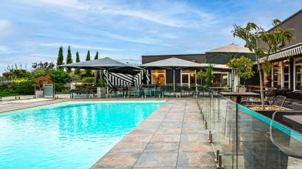 Piscine extérieure du Novotel Reims Tinqueux entourée de terrasses aménagées avec parasols, végétation soignée et baies vitrées du restaurant, sous un ciel bleu légèrement nuageux.
