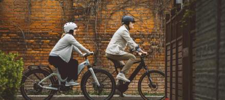 Deux cyclistes portant des casques roulent sur des vélos électriques le long d’un mur en briques avec végétation, Fayet Hospitality.
