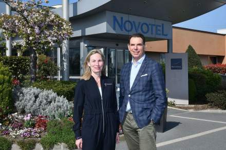 Deux personnes posant devant l’entrée d’un établissement Novotel, sous un ciel ensoleillé.