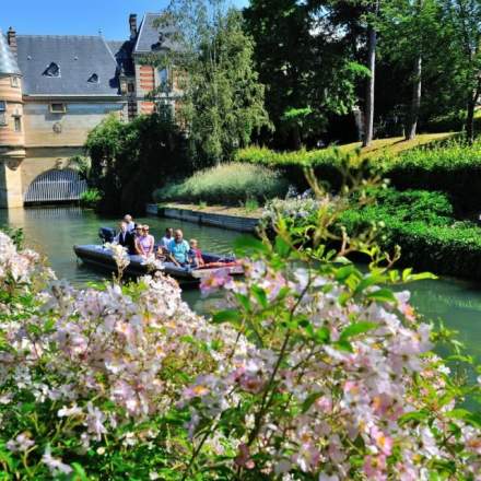 Balade en barque sur une rivière bordée de fleurs et verdure, avec vue sur un bâtiment historique – Fayet Hospitality