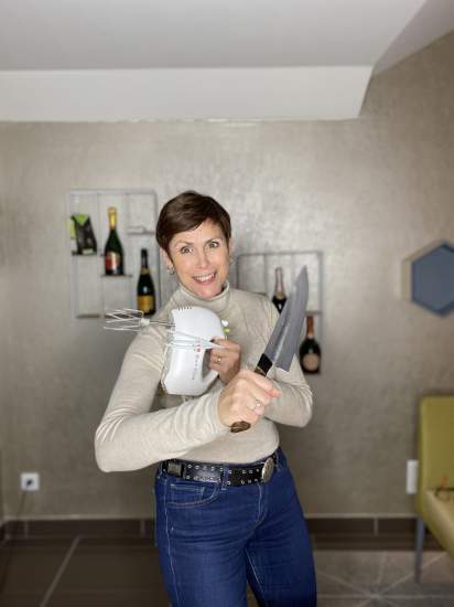 Personne tenant un batteur électrique et un grand couteau dans une cuisine moderne, arrière-plan avec bouteilles de champagne sur étagères murales – Fayet Hospitality