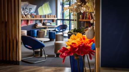 Espace détente de l’Hôtel Ibis Budget Pontault-Combault, avec fauteuils colorés, petites tables, étagères remplies de livres et décoration florale.