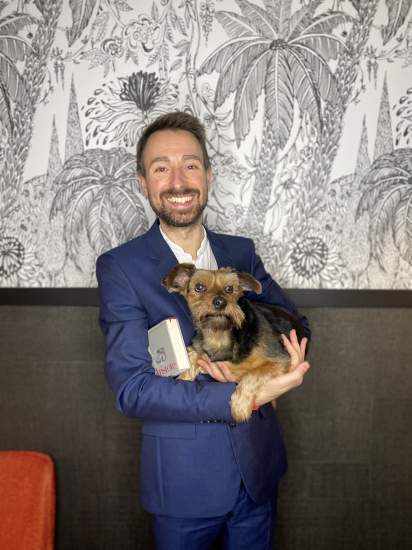 Petit chien brun et noir tenu dans les bras d’une personne portant un costume bleu, fond décoratif avec motifs floraux en noir et blanc – Fayet Hospitality