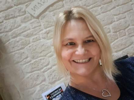 Cheveux blonds lissés avec collier argenté en forme de cœur, fond en pierre claire avec plaque murale visible – Fayet Hospitality
