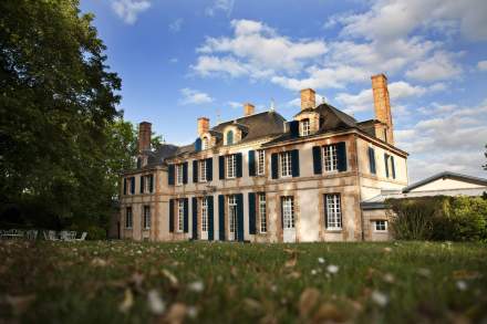 Façade d’un élégant manoir en pierre avec volets bleus et cheminées, entouré de verdure sous un ciel bleu parsemé de nuages – Fayet Hospitality
