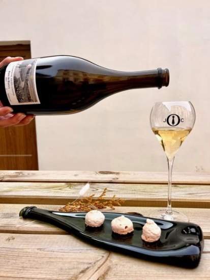 Bouteille de vin en train d’être servie dans un verre à pied marqué JC, posés sur une table en bois avec assiette noire contenant trois bouchées gastronomiques - Fayet Hospitality