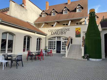 Cour intérieure avec tables et chaises colorées devant la façade Champagne Dauby, grande bouteille décorative verte, Fayet Hospitality