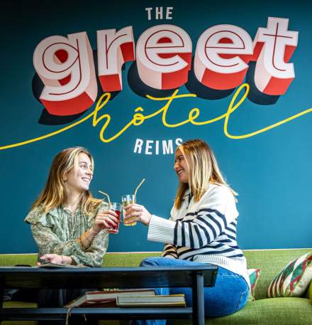 Deux clientes trinquant autour d’une boisson dans l’espace lounge du greet hôtel Reims Tinqueux, devant une grande fresque colorée du logo de l’établissement.