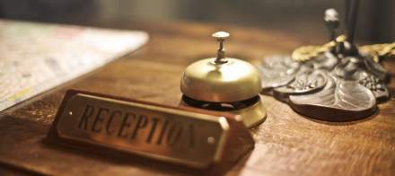 Cloche de réception en laiton posée sur un comptoir en bois avec plaque indiquant “Reception” en arrière-plan – Fayet Hospitality
