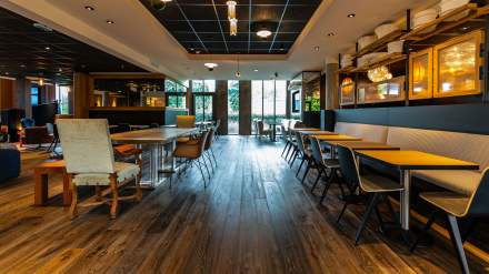 Salle de restauration moderne et spacieuse avec parquet en bois, tables alignées, chaises contemporaines et grande baie vitrée apportant de la lumière naturelle.