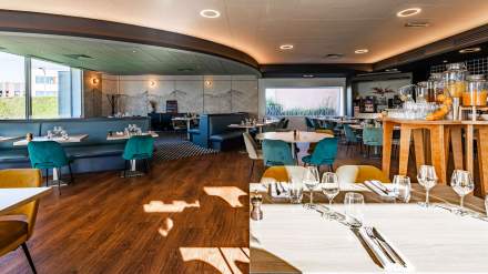 Salle de restaurant lumineuse et moderne, avec mobilier coloré, tables dressées et larges baies vitrées offrant une vue extérieure.