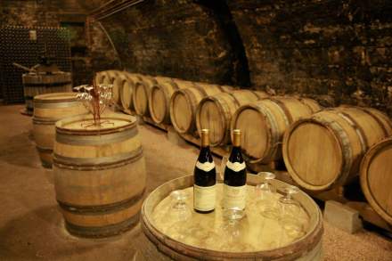 Bouteilles de vin posées sur un tonneau en bois dans une cave voûtée avec rangée de fûts en arrière-plan, ambiance authentique et traditionnelle - Fayet Hospitality