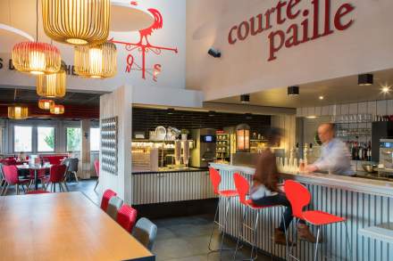Bar du restaurant Courtepaille de l’Hôtel Ibis Reims Tinqueux, avec tabourets rouges, luminaires en bois, comptoir moderne et espace de restauration en arrière-plan.