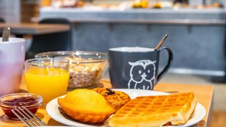 Petit-déjeuner de l’Hôtel Ibis Budget Reims Thillois, composé de gaufre, madeleine, tartelette, confiture, jus d’orange, céréales et boisson chaude servie dans une tasse illustrée.