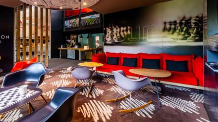 Espace lounge de l’Hôtel Ibis Reims Tinqueux, avec canapé rouge, fauteuils design, tables basses et décoration murale moderne.