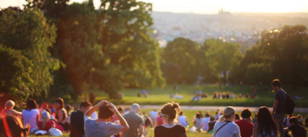 Personnes assises sur l’herbe dans un parc verdoyant au coucher du soleil, avec vue sur la ville en arrière-plan, Fayet Hospitality.