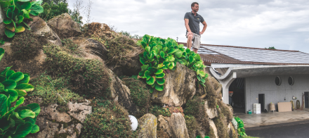 Personne debout sur un rocher couvert de végétation devant un bâtiment moderne avec toit équipé de panneaux solaires – Fayet Hospitality