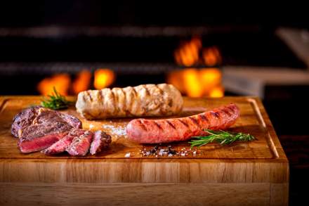 Assortiment de viandes grillées sur planche en bois, comprenant steak émincé, saucisse grillée et pomme de terre au four, avec herbes aromatiques devant un feu de cuisson.