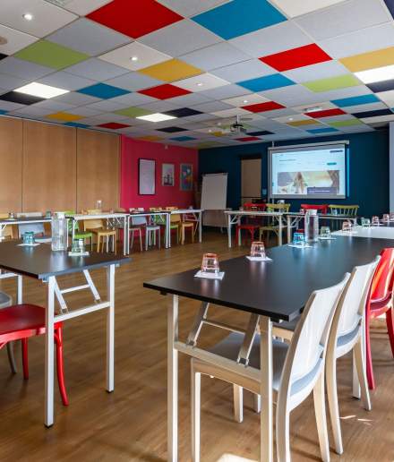 Salle de réunion spacieuse et colorée, aménagée avec tables modernes, chaises variées et écran de projection allumé.