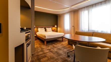 Suite d’hôtel lumineuse avec un grand lit double, un canapé d’angle beige, une table ovale avec chaise, et un coin kitchenette, le tout dans une ambiance moderne et chaleureuse.