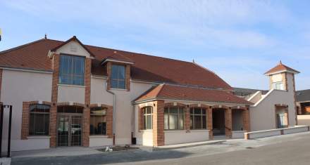 Bâtiment moderne avec murs clairs et encadrements en briques rouges, toit en tuiles et tour latérale, sous ciel bleu – Fayet Hospitality