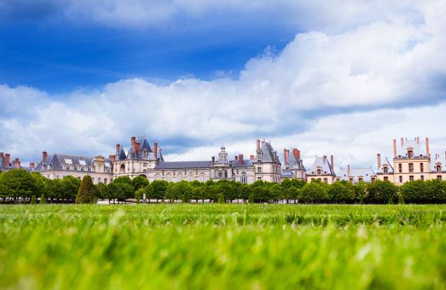 Grand château historique entouré d’un vaste parc verdoyant sous un ciel bleu parsemé de nuages.