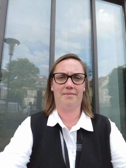 Personne vêtue d’une chemise blanche et d’un gilet noir assise devant une façade vitrée avec reflet d’arbres et bâtiments – Fayet Hospitality