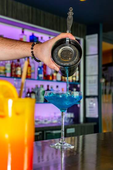 Un barman verse un cocktail bleu depuis un shaker dans un verre à pied, avec un autre cocktail coloré au premier plan et un bar illuminé en arrière-plan.