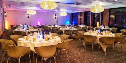 Salle de réception élégante avec tables rondes dressées, chaises design et éclairages décoratifs chaleureux.