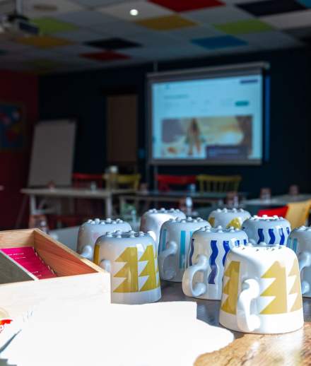 Salle de réunion colorée avec vidéoprojecteur allumé, matériel de travail et tasses décorées disposées sur une table.
