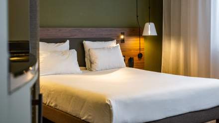 Chambre d’hôtel moderne avec un grand lit double, tête de lit en bois, lampes suspendues et rideaux clairs laissant entrer une lumière douce.