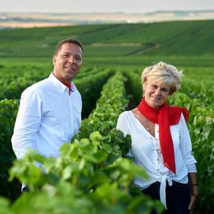 Deux personnes en chemises blanches avec foulards rouges se tenant dans des rangées de vignes verdoyantes sous ciel clair, Fayet Hospitality.
