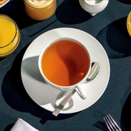 Tasse de thé chaud sur soucoupe blanche avec cuillère, entourée de verres de jus et d’éléments de petit-déjeuner – Fayet Hospitality