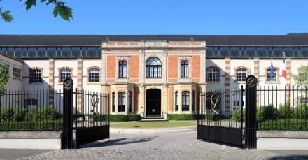 Façade élégante d’un bâtiment historique en briques avec portail en fer forgé ouvert, drapeau français visible à droite – Fayet Hospitality