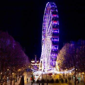 Grande roue illuminée en violet et blanc la nuit, attraction festive au cœur d’une ville animée - Fayet Hospitality