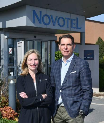 Entrée d’un hôtel Novotel avec enseigne visible, deux personnes en tenue professionnelle posant devant le bâtiment, végétation décorative et signalétique à l’extérieur – Fayet Hospitality