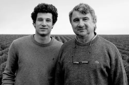 Deux personnes debout dans un vignoble, portant des vêtements sobres, avec rangs de vigne en arrière-plan – Fayet Hospitality