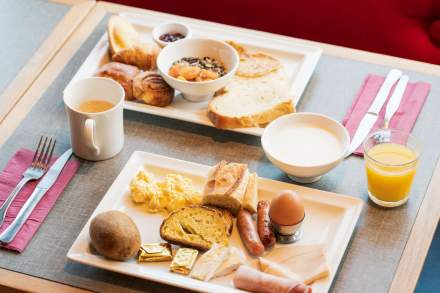 Petit-déjeuner complet de l’Hôtel Ibis Reims Tinqueux, avec œuf, charcuterie, fromage, pain grillé, viennoiseries, fruits, céréales, café et jus d’orange servis sur plateau.