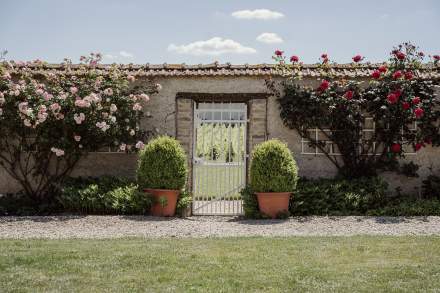 Portail en fer forgé entouré de rosiers grimpants et de deux buissons taillés dans des pots en terre cuite, devant un mur en pierre sous ciel ensoleillé - Fayet Hospitality