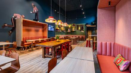 Salle de restauration du greet hôtel Reims Tinqueux, avec banquettes colorées, grandes tables en bois, décor artistique mural et luminaires design suspendus.